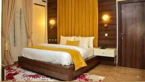 A bedroom with a wooden headboard, a bedside table, and a bed with white bedding and a yellow throw. - RKD SUITES Asaba (Asaba)