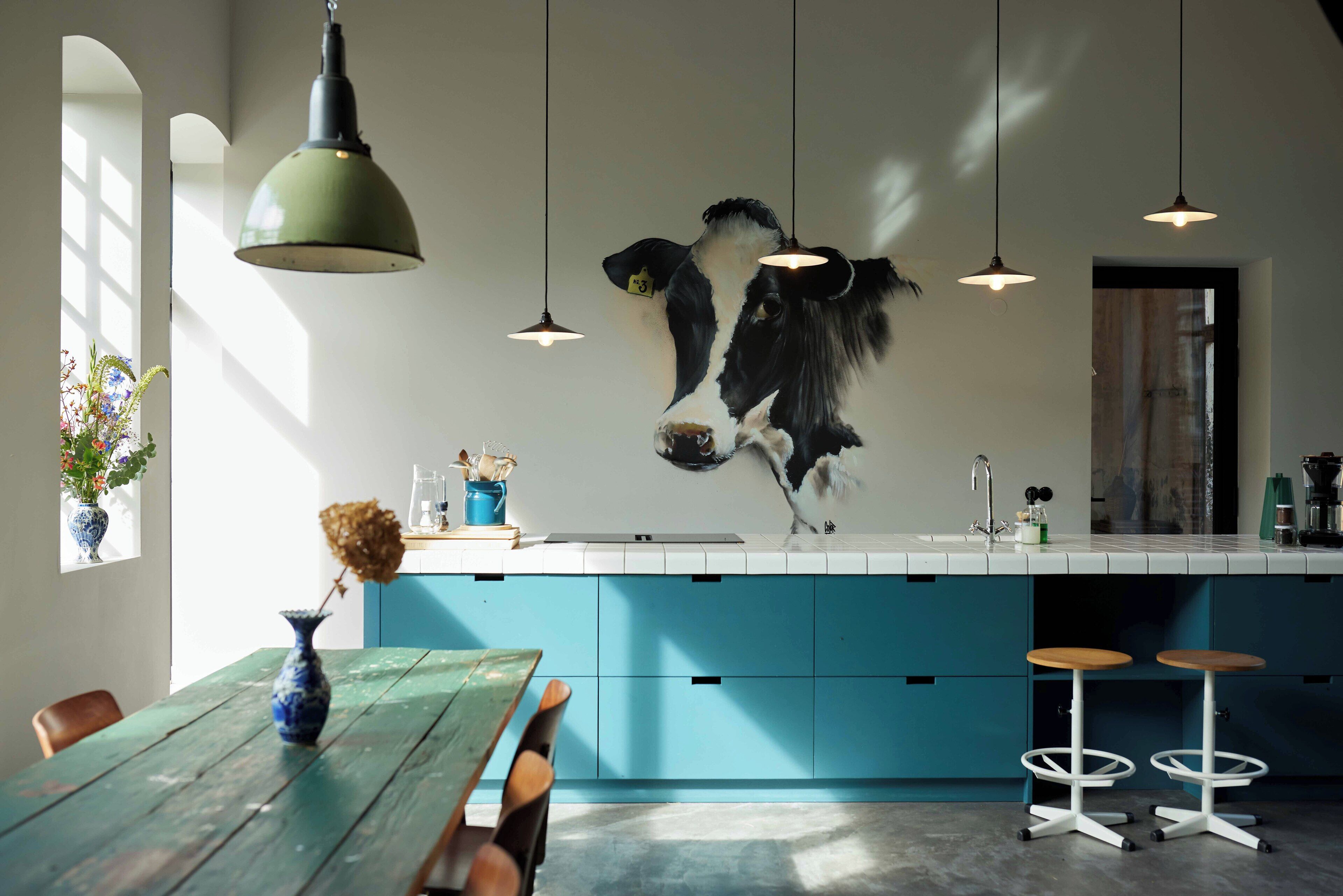A modern kitchen with blue cabinets, a hanging light fixture, and a large wall-mounted cow decoration.