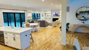 A modern living room with a fireplace, a dining area with a white table and chairs, and a kitchen with white cabinets and a built-in oven.