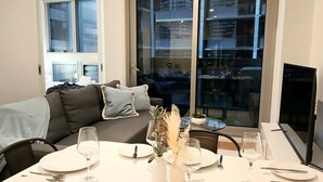 Ruokailutila, jossa on neljälle katettu ruokapöytä, tyynyillä varustettu sohva ja suuri ikkuna, josta avautuu näkymä kaupunkimaisemaan.