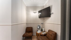 Una habitación pequeña y bien iluminada con dos sillones de cuero marrón, un televisor de pantalla plana montado en la pared, una mesita con un ventilador y una taza, y una cortina a la derecha.