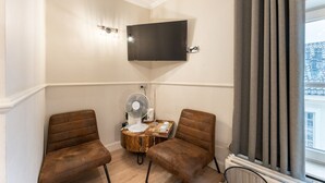 Habitación lateral con dos sillas marrones, una mesita de madera, un ventilador blanco y un televisor de pantalla plana montado.