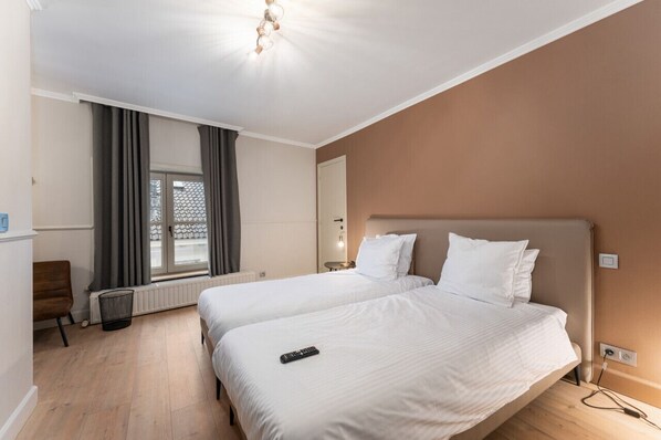 Une chambre d’hôtel avec deux lits, une fenêtre avec des rideaux, un radiateur, une table de chevet et un luminaire fixé au mur.