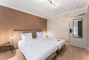Un lit double avec une literie blanche, une table de chevet avec une lampe et une porte dotée d’un miroir.