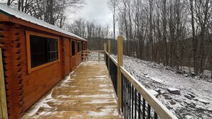 A wooden cabin with a wooden deck and a metal railing, surrounded by a snowy landscape and leafless trees. - The Cozy Cabin: Mountain Escape at Snowshoe, WV (Slaty Fork)