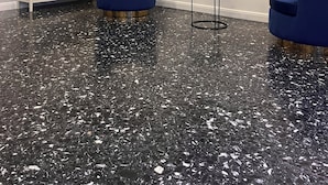 Et værelse med et sort marmoreret gulv, hvide vægge med paneler, et fladskærms-tv monteret på væggen og to blå stole med guld detaljer.