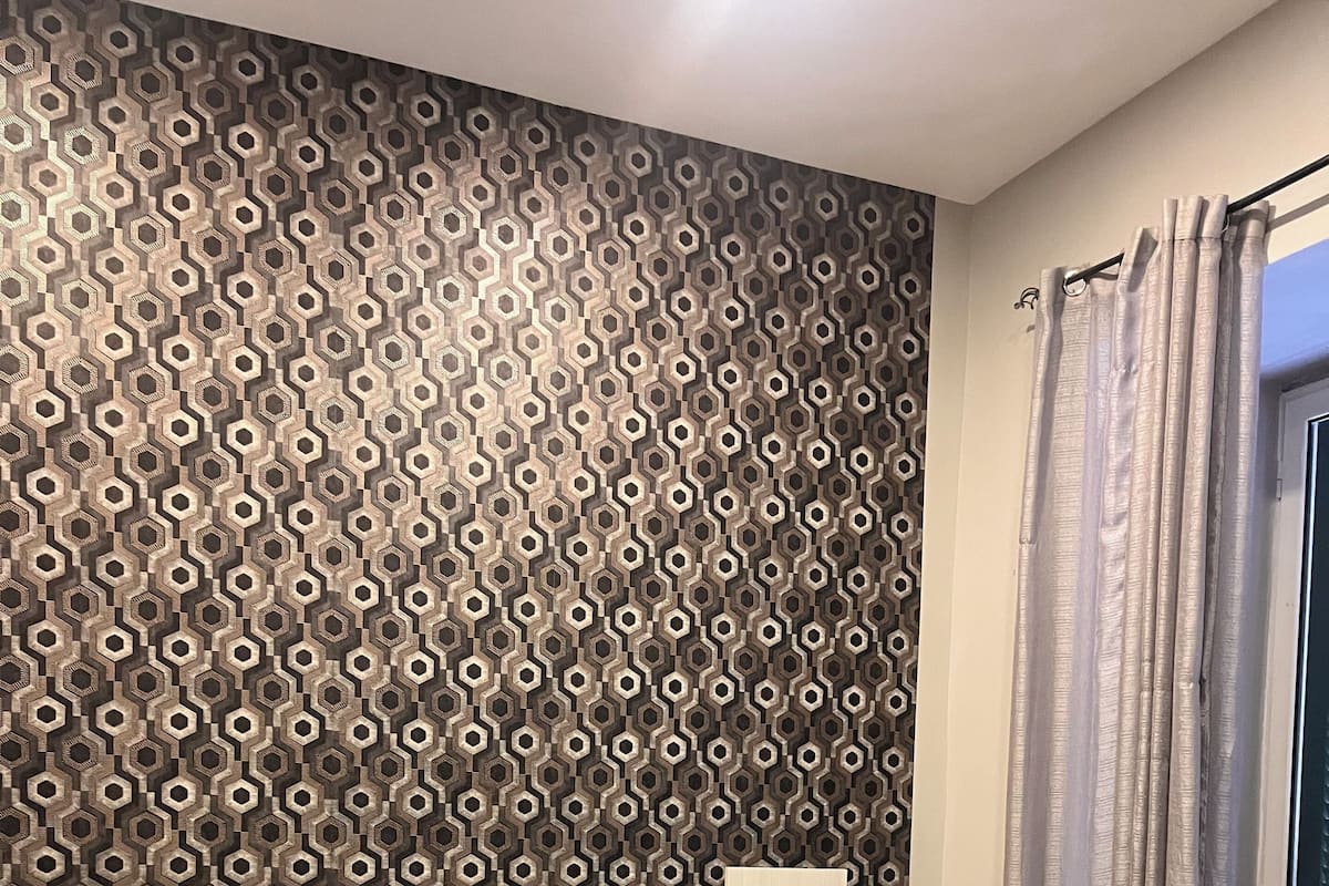 A hotel room with a patterned wall, a bed with a dark headboard, a bedside table with a lamp, and a window with curtains.