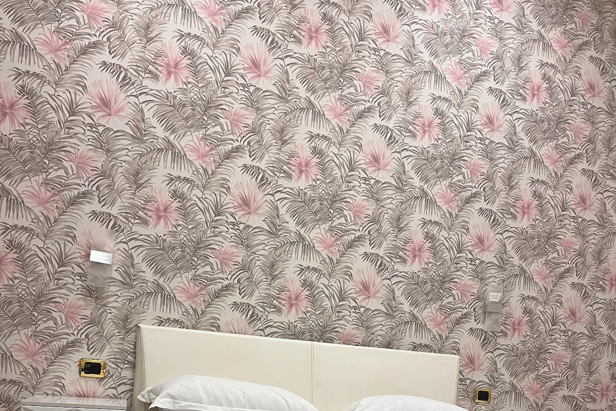 A neatly made bed with white linens and a folded towel, a white nightstand with a lamp, and a wall with a tropical leaf pattern.