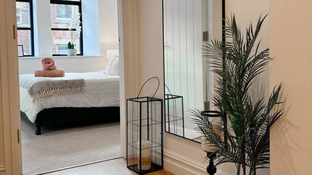 A hallway with a large mirror, a potted plant, and a bed in the adjacent room. - Modern Leeds Central Stay -2 min walk from station (Leeds)