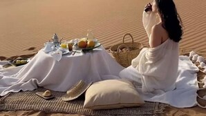 A desert scene with a person sitting on a blanket, a picnic setup, and sand dunes in the background.