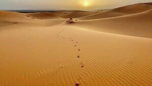 A desert landscape with sand dunes and a vehicle's tracks leading towards the sun.