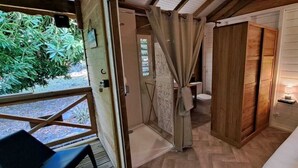Een kamer met een houten bed, een kleine badkamer met toilet en douche, en een raam met uitzicht op groen.