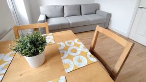 A dining area with a wooden table, two chairs, a potted plant, and a grey sofa in the background.