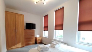 A bedroom with a bed, a TV, a wardrobe, and two windows with blinds.