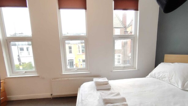 A hotel room with a large bed, three windows, a radiator, and a view of buildings outside.