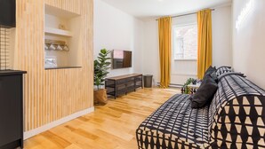 A modern living room with a wooden partition, a sofa with patterned cushions, a TV, a small table, and a potted plant. - HOMA-Comfort&Style-2BR-Spacious-Natural Light-FeelHome-A/C-SelfCheckIn-Metro (Montréal)