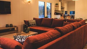 Ein modernes Wohnzimmer mit einem Ecksofa, einem Flachbildfernseher und einem Essbereich, im Hintergrund ist eine Küche zu sehen.