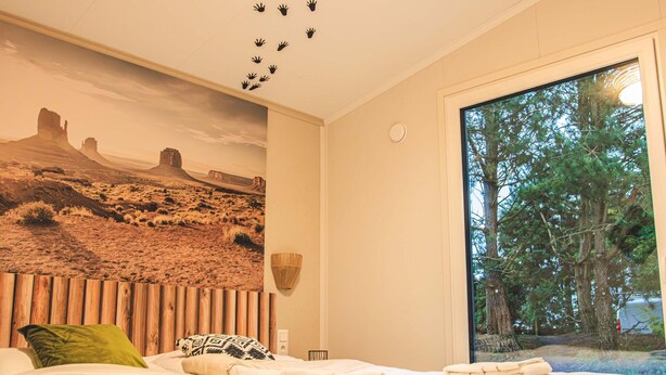 Ein Schlafzimmer mit Wüstenwandmalerei, einem großen Fenster mit Blick auf einen Wald und einem Bett mit ordentlich gefalteten Handtüchern.