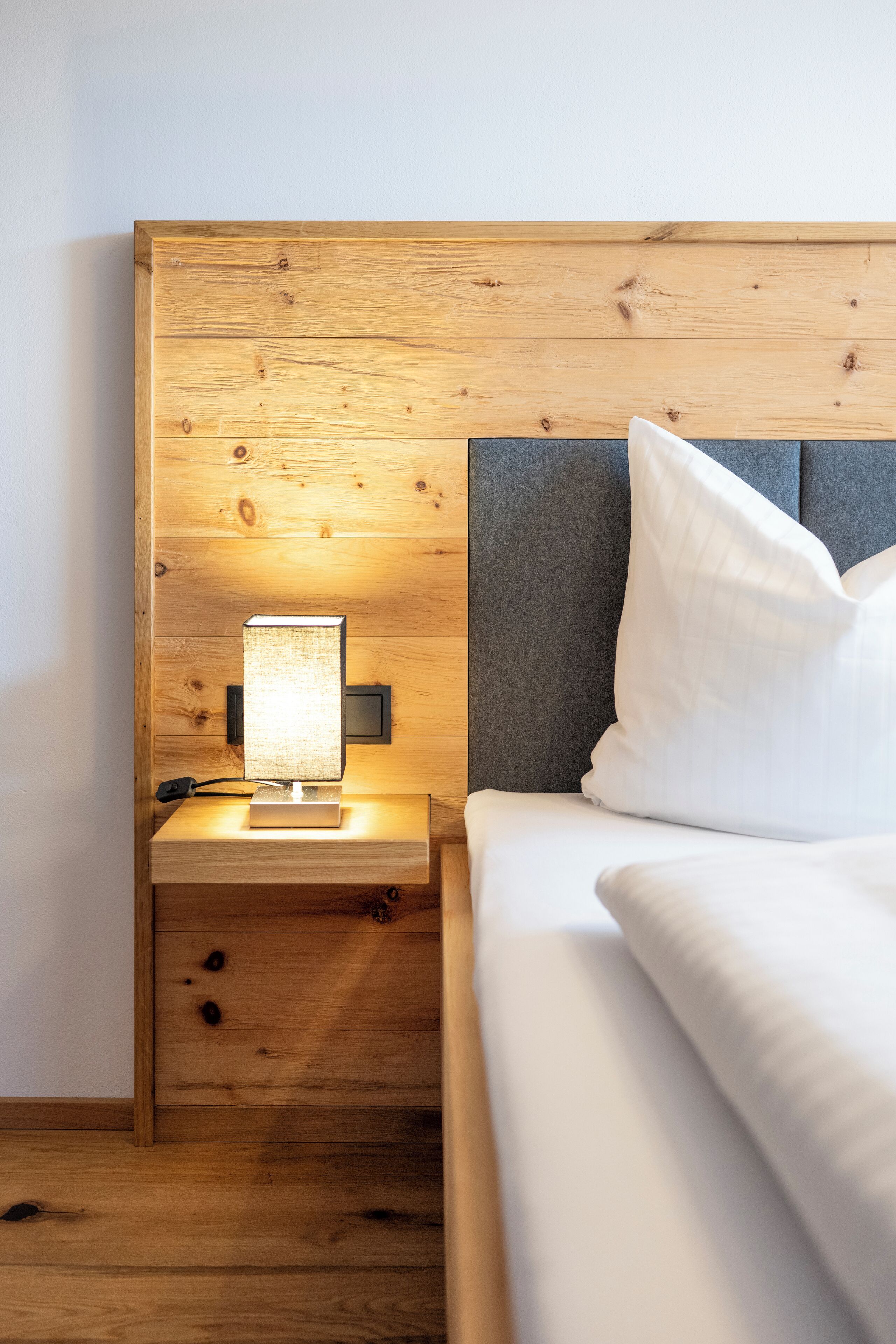 Een net opgemaakt bed met witte lakens en een kussen, een nachtkastje met een lamp, en een houten hoofdbord.