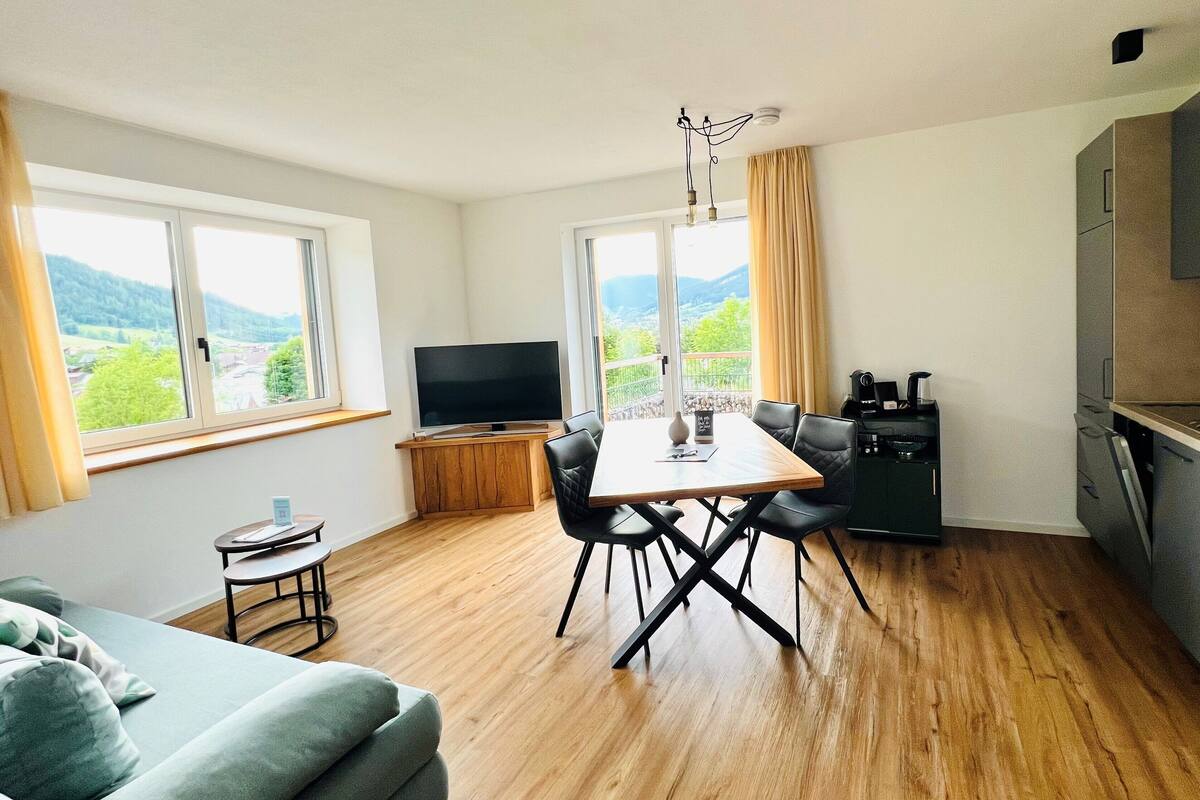 A modern apartment with a dining area, kitchen, and a large window offering a scenic view.