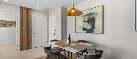 Un comedor moderno con una mesa blanca, sillas oscuras, una lámpara colgante y un cuadro de un paisaje urbano en la pared.