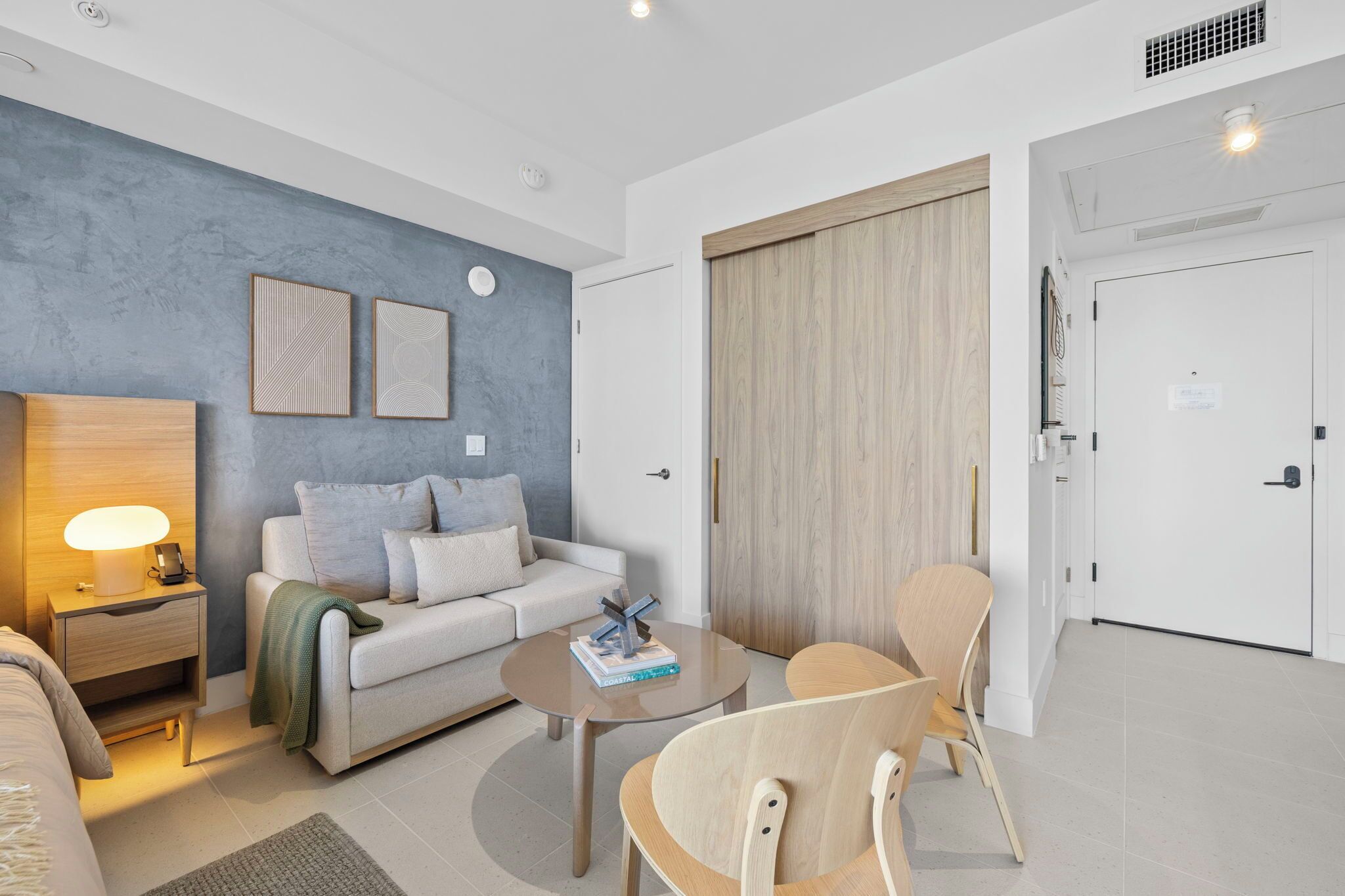 A modern living room with a sofa, coffee table, and chairs. There is a wooden nightstand with a lamp, a wall with framed artwork, and a sliding door.