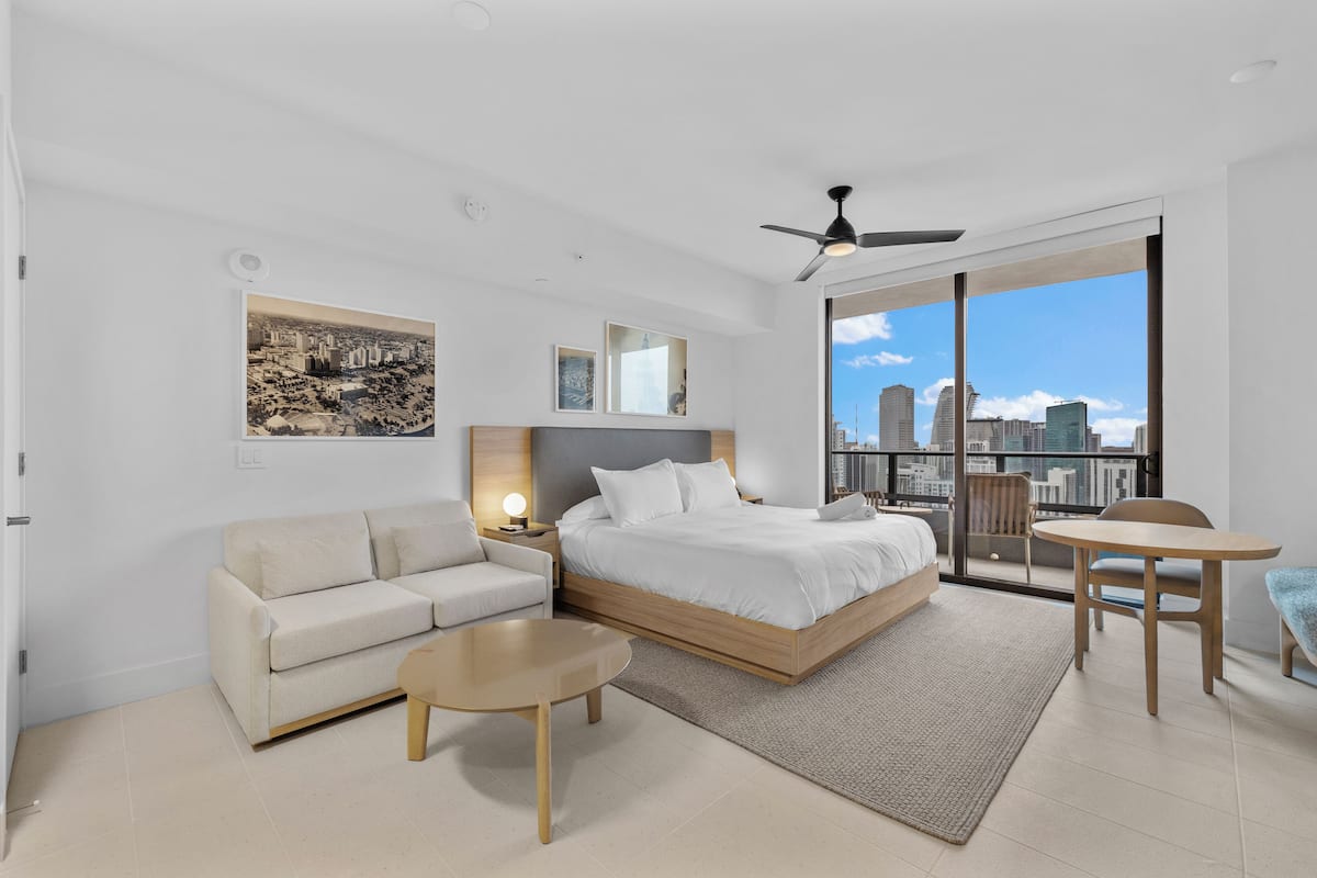 A modern bedroom with a large bed, a sofa, a small table, and a balcony with a city view.