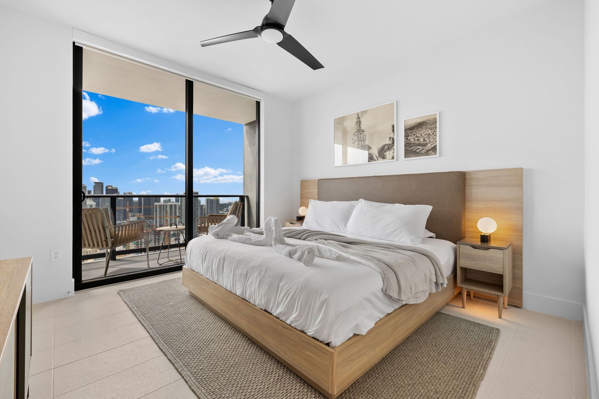 Une chambre moderne avec un grand lit, un ventilateur de plafond, une vue sur la ville par une porte coulissante et deux tableaux encadrés au mur.