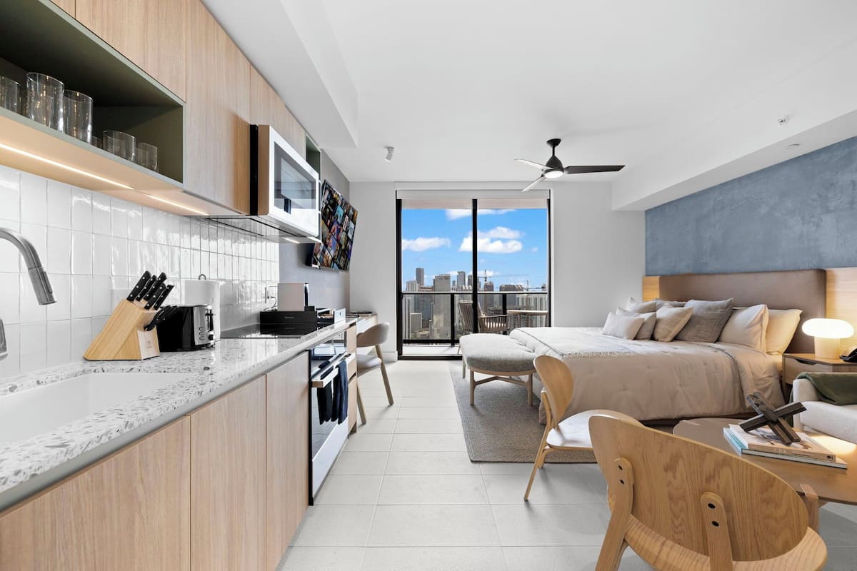A modern apartment with a kitchen, dining area, and bedroom. It features a city view, a ceiling fan, and a large bed.
