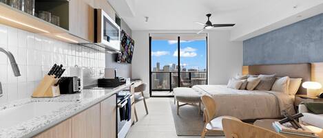 A modern apartment with a kitchen, dining area, and bedroom. It features a city view, a ceiling fan, and a large bed.