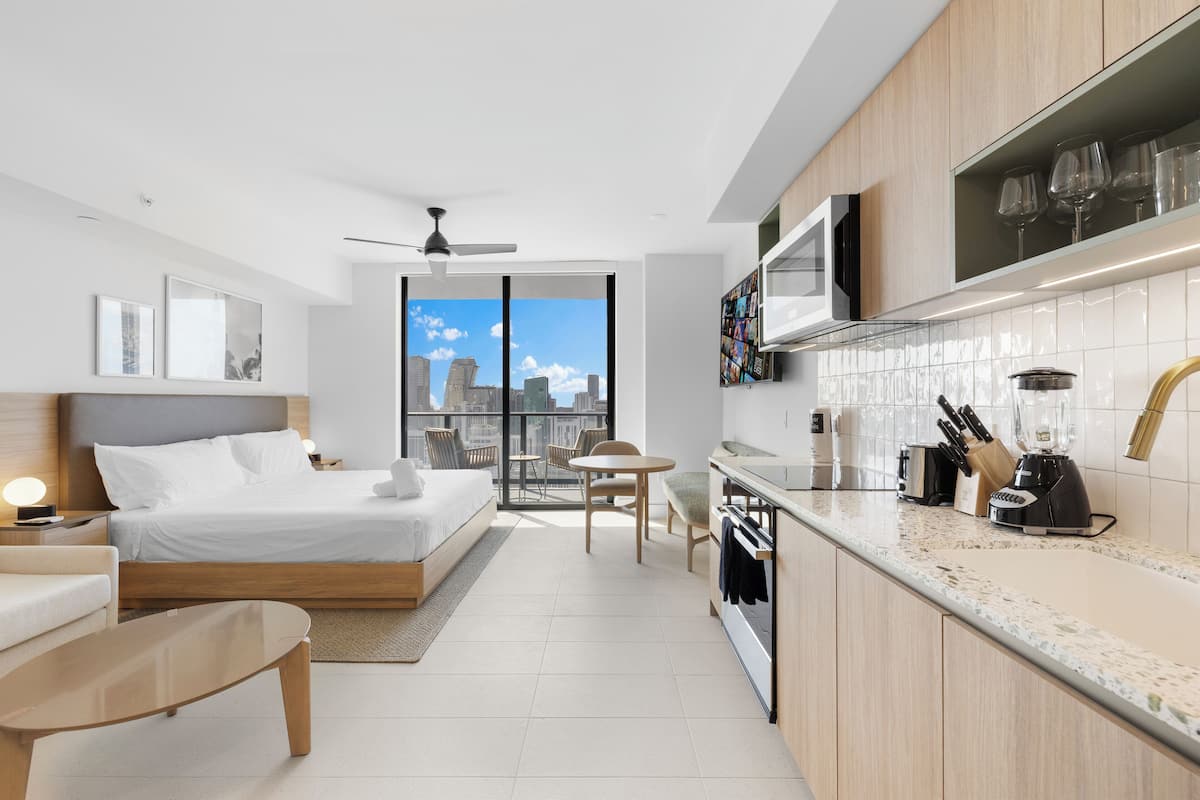 A modern hotel room with a bed, a sitting area, a kitchen with a dining table, and a view of the city.