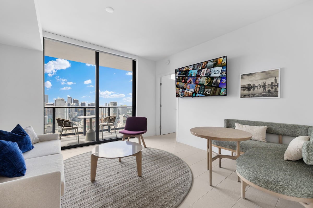 A modern living room with a large window, a city view, a round table, a sofa, and a wall art piece.