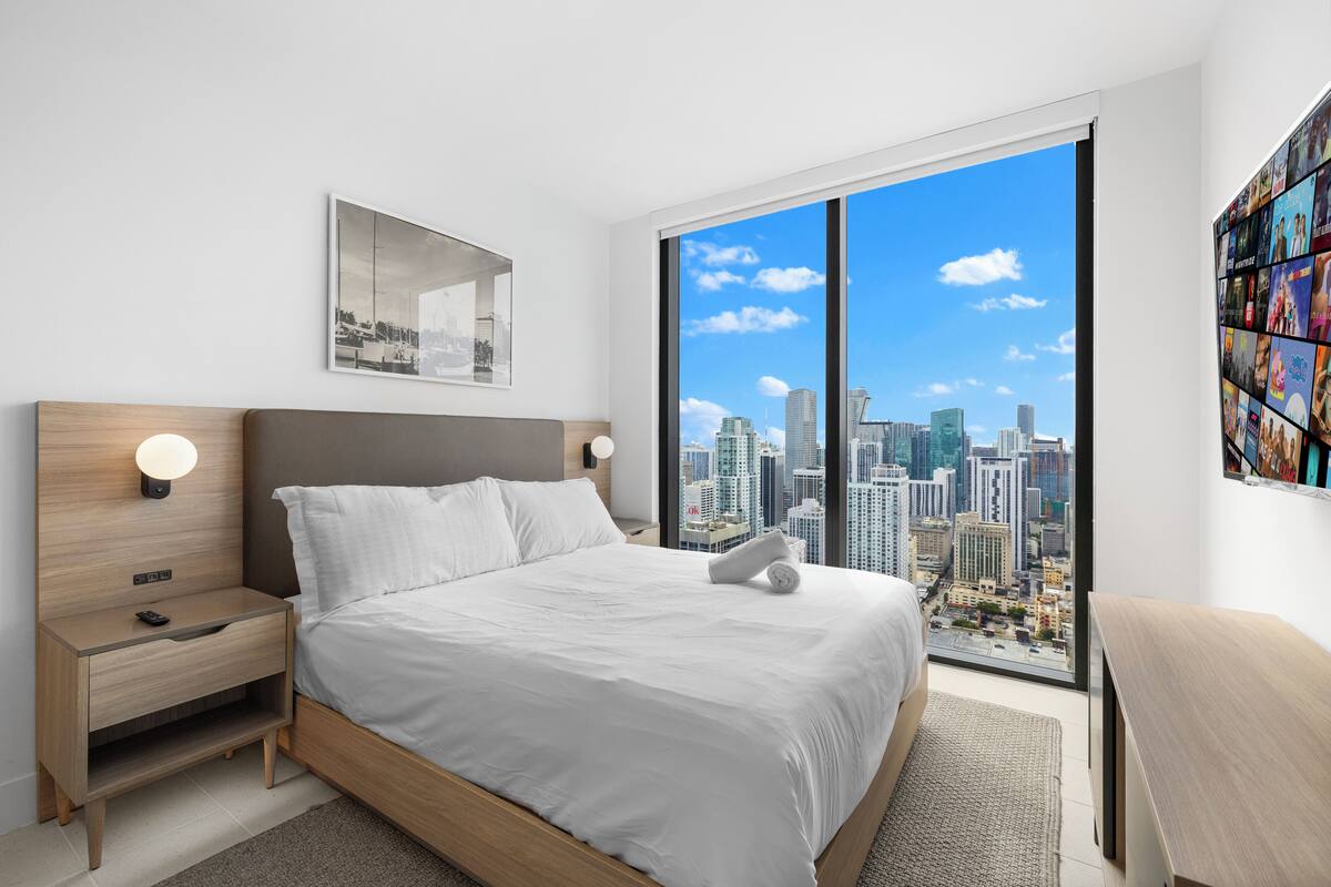 A modern bedroom with a large bed, a nightstand, a wall-mounted TV, and a city view through large windows.