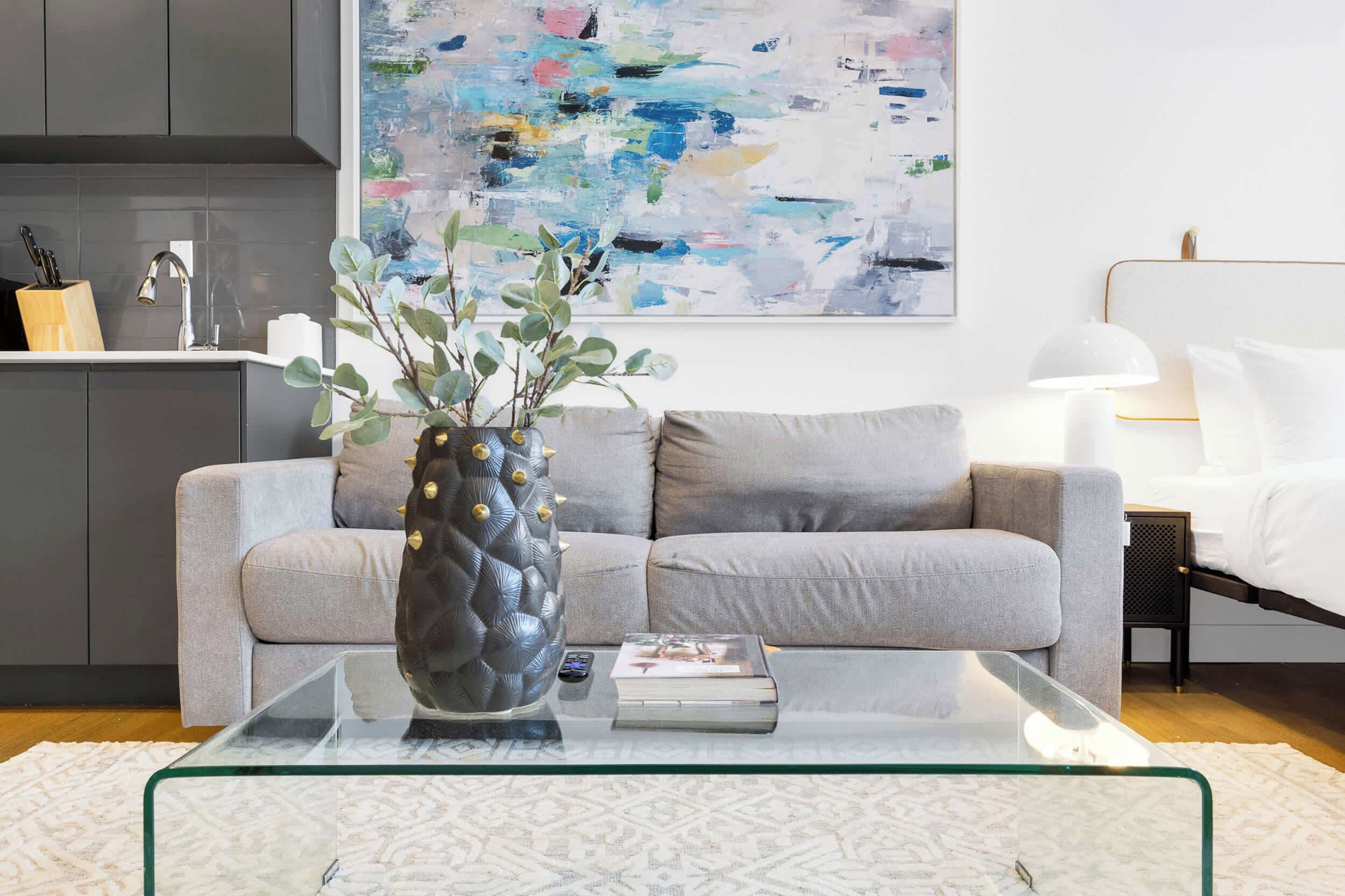 A modern living room with a glass coffee table, a grey sofa, a white rug, a painting on the wall, and a kitchen area in the background.
