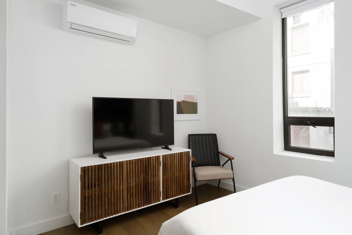 Una habitación de hotel moderna con una televisión de pantalla plana sobre un mueble de madera, una cama individual, una silla y un ventanal con vistas a edificios.