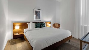 A modern hotel room with a wooden bed, two bedside lamps, a framed artwork on the wall, and a chair by the window. - Modern One-Bedroom|Downtown Steps from Centre Bell (Montréal)