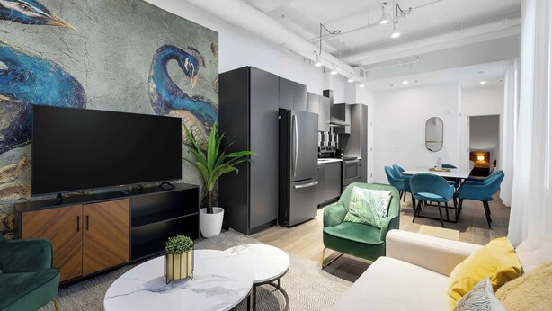 A modern living room with a flat-screen TV, a dining area with blue chairs, and a kitchen with black cabinets. - Modern One-Bedroom|Downtown Steps from Centre Bell (Montréal)