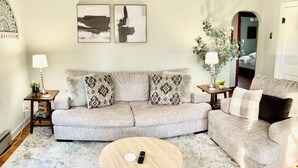 A living room with a grey sofa, a round coffee table, and a side chair. There are two wall-mounted paintings, a lamp on a side table, and a small plant. - Stumble-In Guest House- Downtown Location (Hermann)