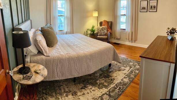 A bedroom with a large bed, a round side table, a lamp, a chair, and a desk with a plant. - Stumble-In Guest House- Downtown Location (Hermann)
