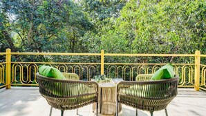 Una terraza con sillas de mimbre y una mesa, rodeada por una barandilla amarilla y abundante vegetación.