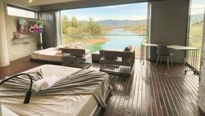 A modern bedroom with a large bed, a desk, and a view of a lake and mountains. - EleMYNT LifeStyle Hotel (Guatapé)