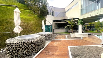 Un área de piscina moderna al aire libre con cubierta de piedra y madera, una palmera y un edificio con fachada de vidrio al fondo.