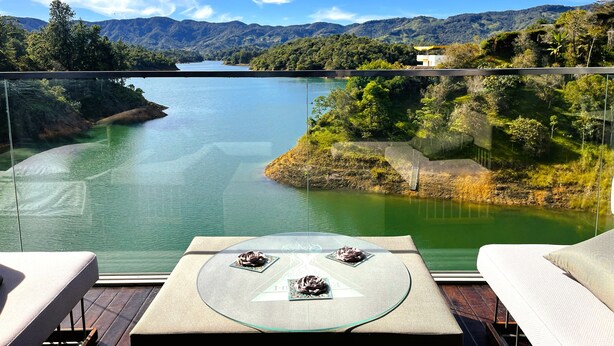 A balcony with a glass table, two chairs, and a view of a lake surrounded by mountains. - EleMYNT LifeStyle Hotel (Guatapé)