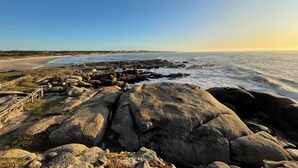 Une côte rocheuse où les vagues s’écrasent contre les rochers, un ciel dégagé et une vue lointaine sur une plage et des bâtiments.