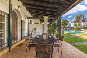 A covered patio with a round table and wicker chairs, a pool in the background, and a view of a residential area.