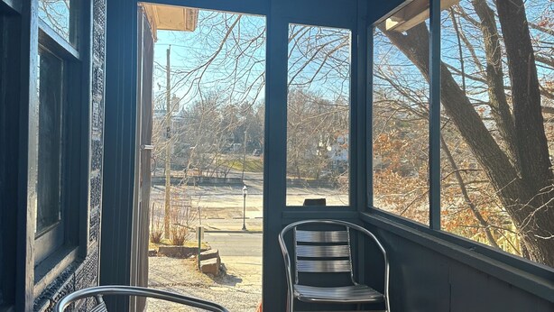 Eine überdachte Veranda mit zwei Metallstühlen und einem Tisch, große Fenster mit Blick auf Bäume und einen Parkplatz.