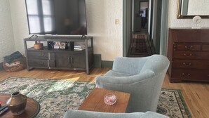 Ein Wohnzimmer mit einem Flachbildfernseher auf einem Schrank, einem grauen Sessel, einem Holztisch und einem Teppich mit Muster.