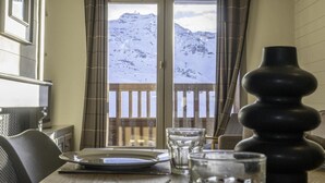 Un coin repas avec une table dressée pour deux personnes, une vue sur des montagnes enneigées à travers une porte vitrée et une balustrade en bois.