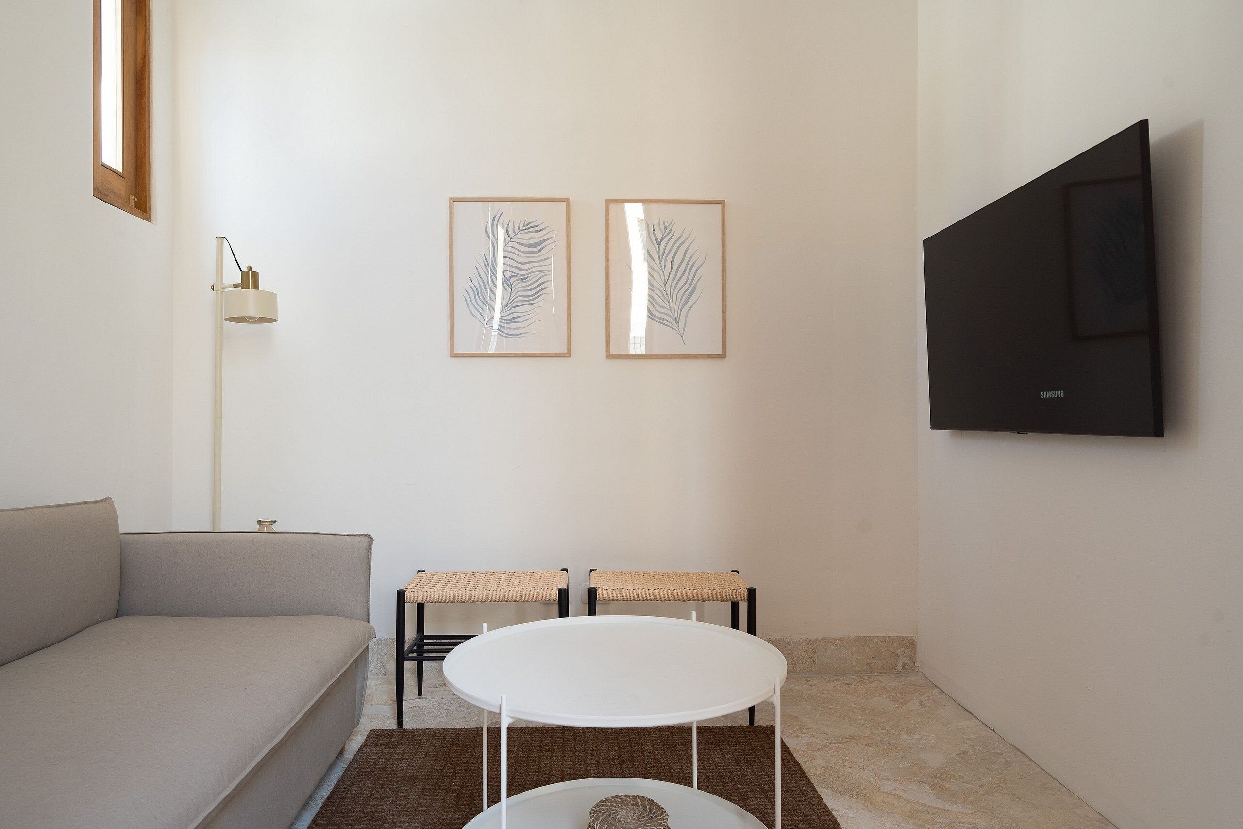 A modern living room with a sofa, a small round table, two framed artworks, a floor lamp, and a mounted TV.