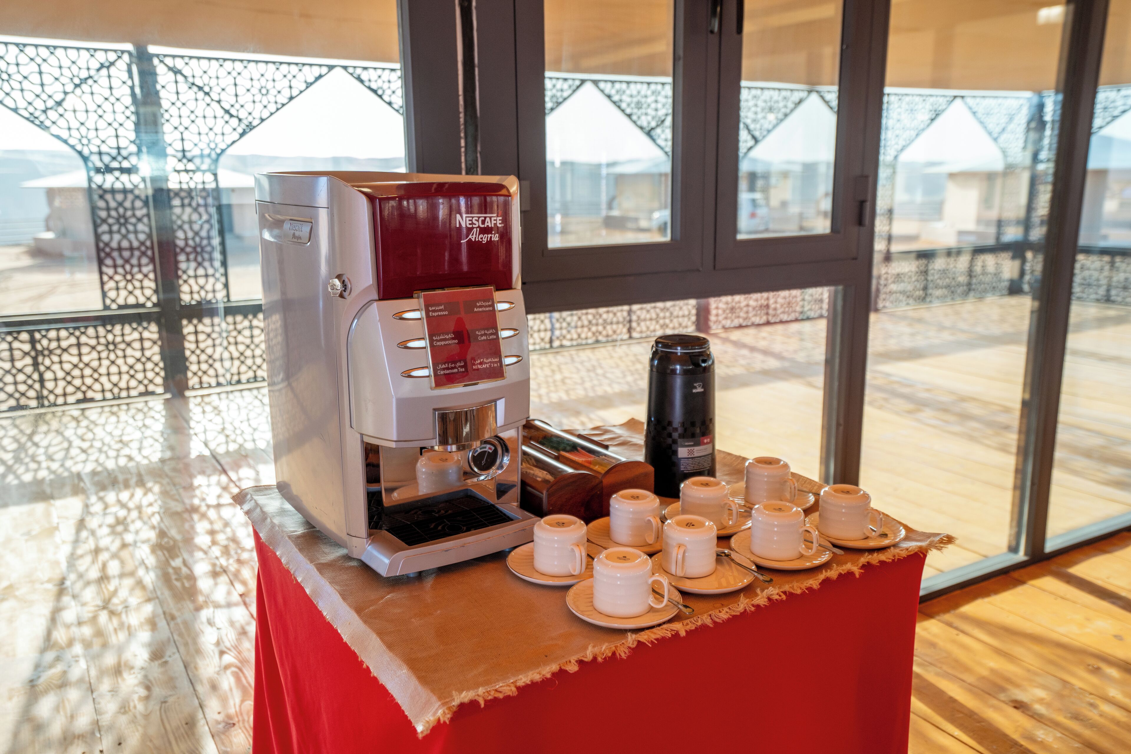 A coffee station with a Nescafe machine, cups, and a thermos on a table in a room with large windows.